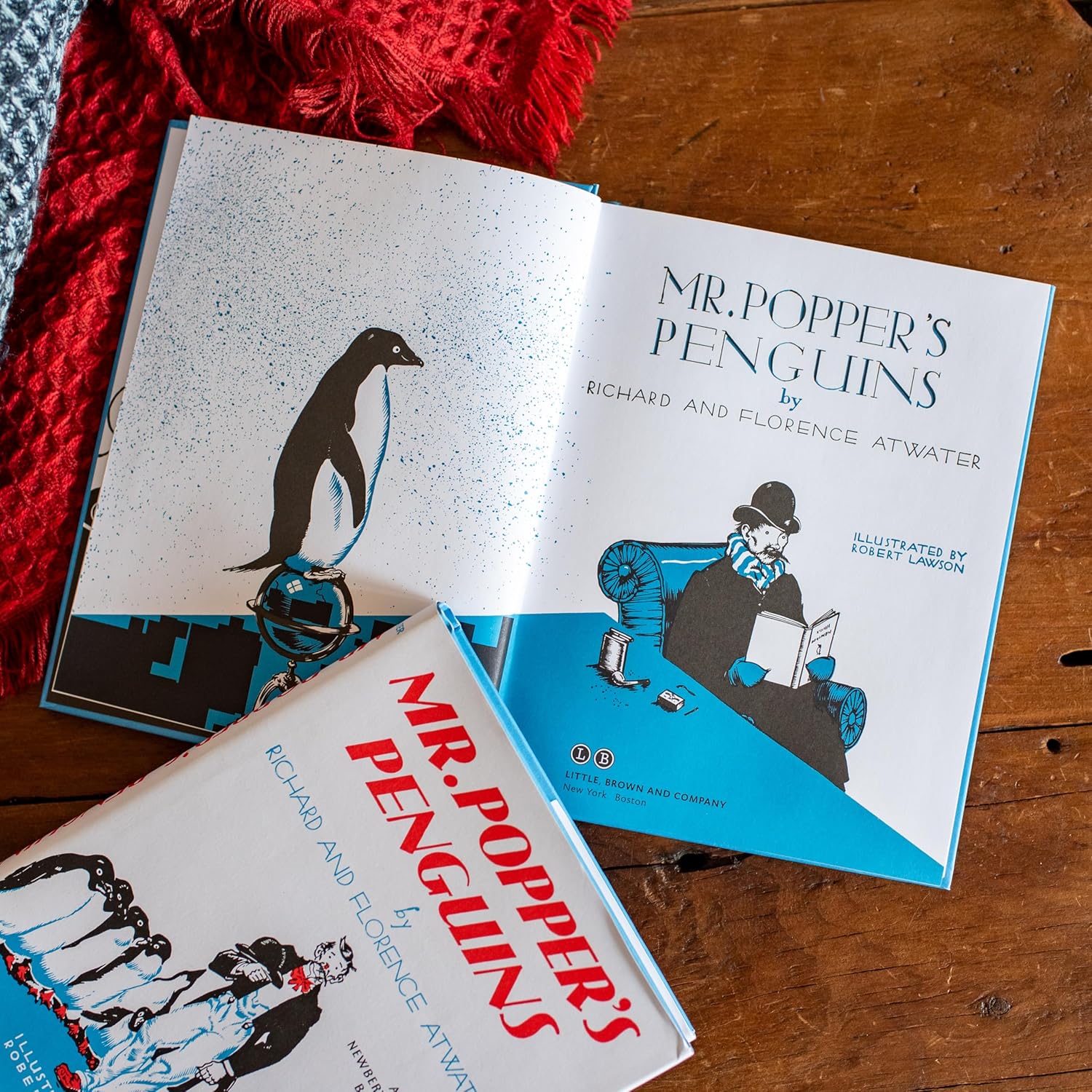 Open book titled 'Mr. Popper's Penguins' on a wooden surface with a red fabric background.